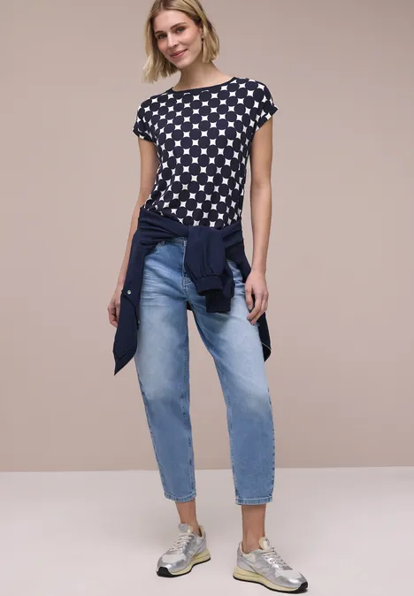 Frau in schwarzem T-Shirt mit weißen Herzen, Jeans, Sneakern und um die Taille gebundener Jacke.